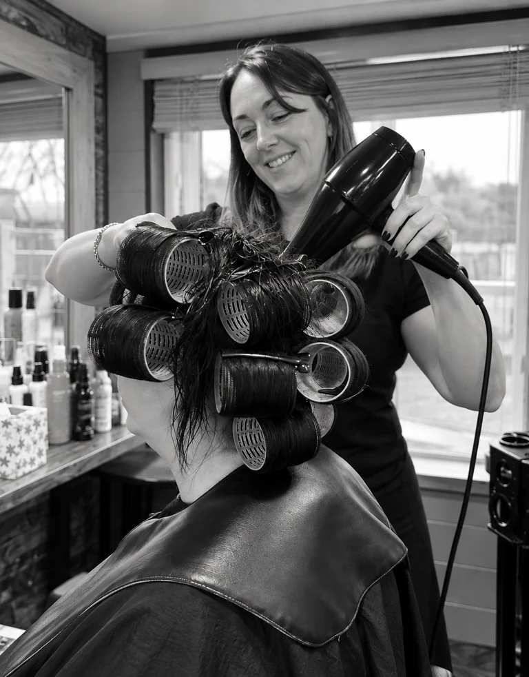 Cerstie smiling as she blow-dries a client’s hair set in rollers, in the sunlit Hair Room
